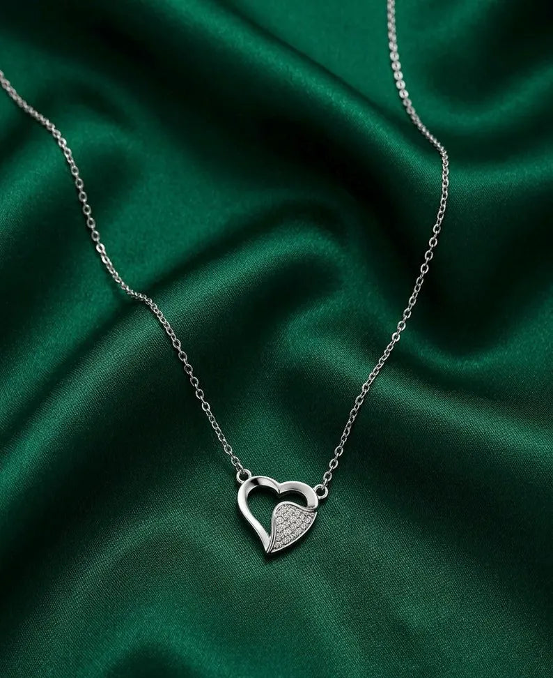 A silver heart pendant necklace with one heart open and one heart filled with small clear stones, displayed on a draped dark green satin fabric.