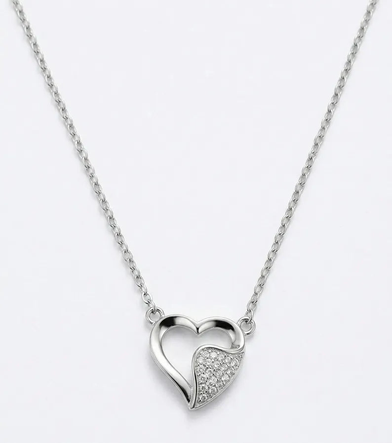 A silver heart pendant necklace on a delicate silver chain, featuring one open heart and one heart paved with small clear stones, set against a plain white background.