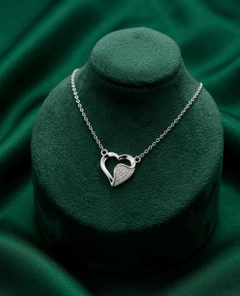 A silver heart pendant necklace with a pavé-set textured heart on a delicate chain, displayed on a dark green velvet jewelry bust against a matching green fabric background.