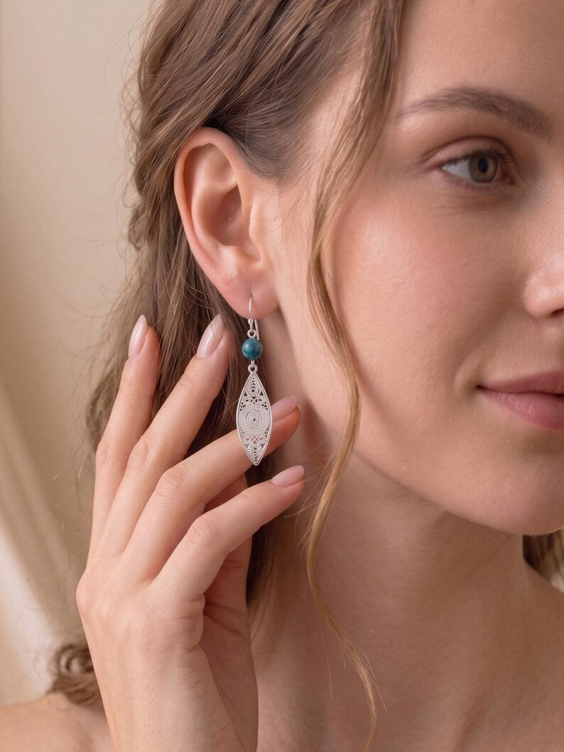 A close-up of a woman wearing a silver filigree dangle earring with a small turquoise bead, touching her ear with her hand.