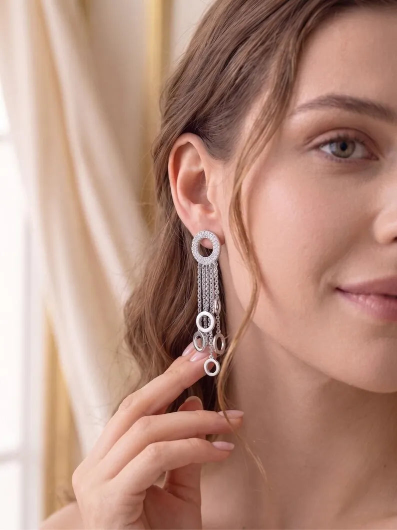 A close-up lifestyle shot of a woman wearing a long, dangling silver earring with multiple circular links and chain details, set against a soft beige curtain background.