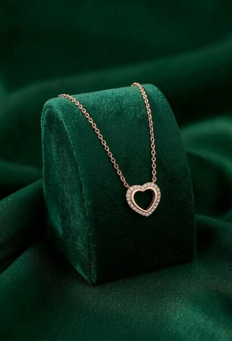 A delicate rose gold heart pendant necklace with a halo setting, resting on a dark green velvet jewelry display stand against a matching green fabric background.