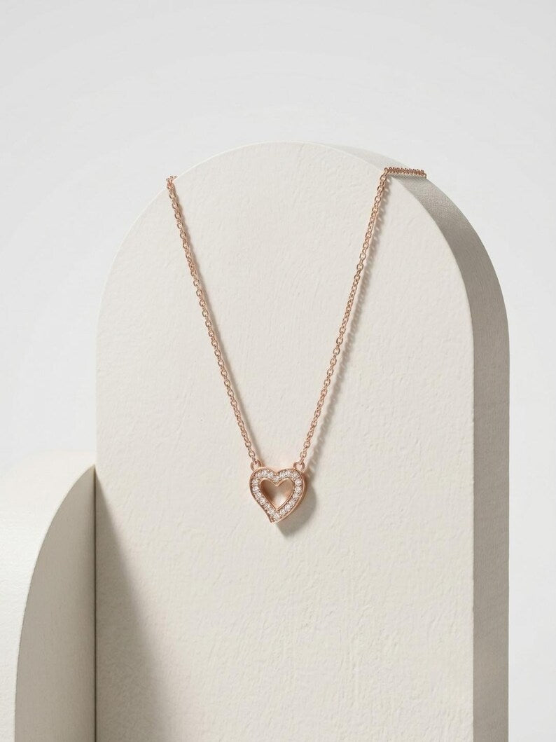 A delicate rose gold chain necklace with a heart-shaped pendant, displayed on a minimalist cream-colored arched stand against a clean white background.
