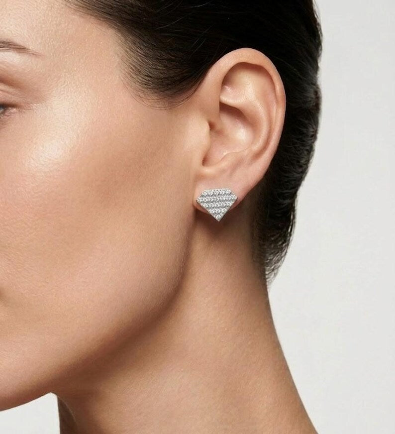 A close-up of a model's ear wearing a small triangular stud earring set with clear stones in a silver-toned metal, against a light gray background.