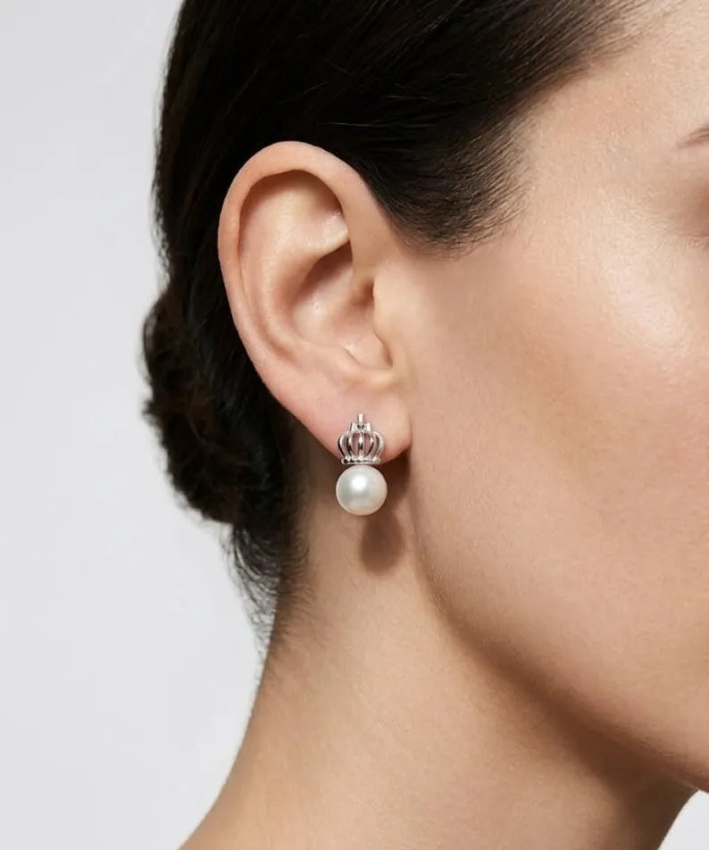 A close-up of a model's ear wearing a silver crown stud earring with a white pearl drop, against a light gray background.