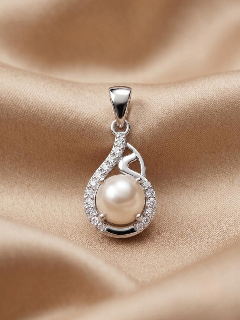 A silver teardrop pendant featuring a central white pearl surrounded by small clear stones, displayed on a draped beige satin fabric background.