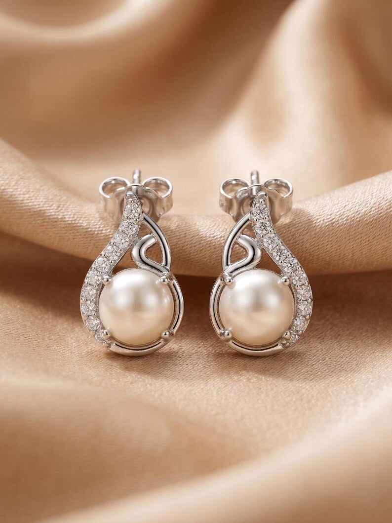 A pair of silver teardrop-shaped earrings with large white pearls and clear pavé accents, displayed on a draped beige satin fabric background.
