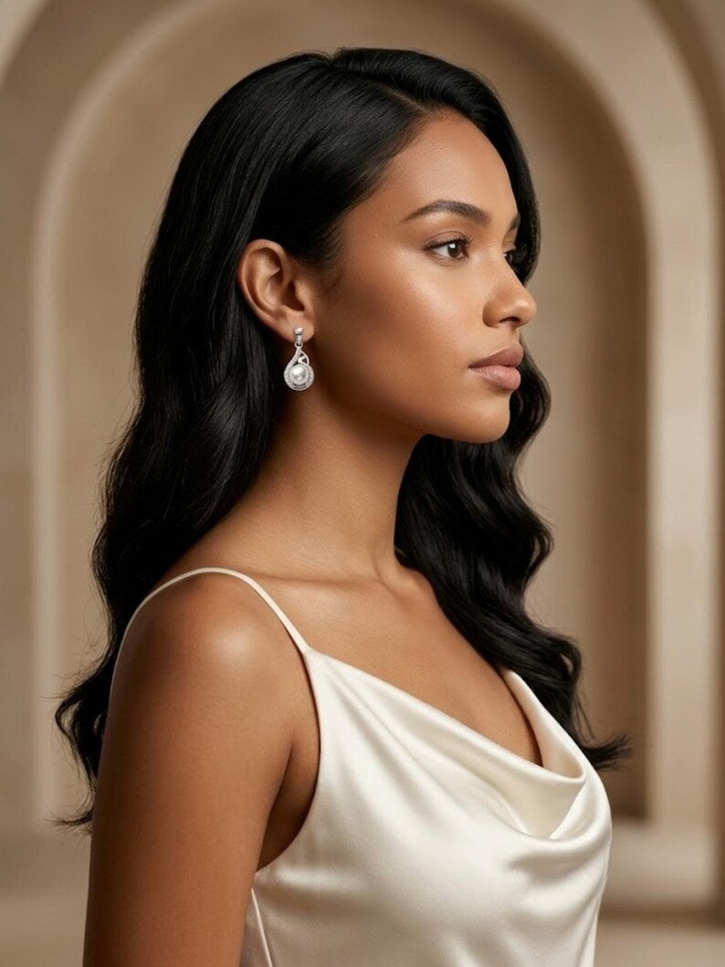 A woman in profile wearing a white satin top and a silver teardrop dangle earring, set against a warm beige arched background.