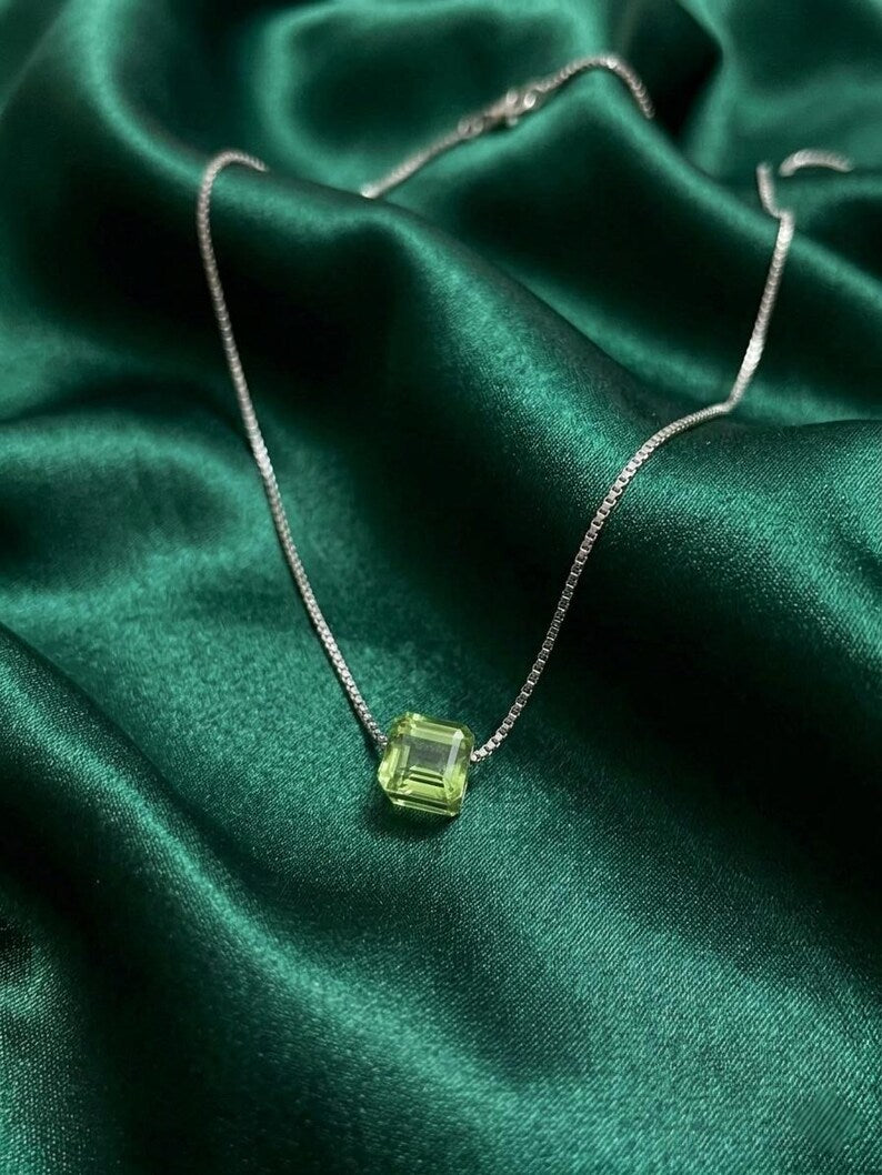A silver chain necklace with a square-cut light green gemstone pendant, displayed on a draped dark green satin fabric background.