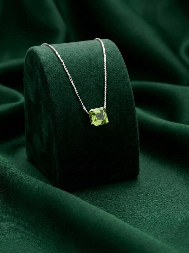 A silver chain necklace with a square green gemstone pendant, displayed on a dark green velvet jewelry stand against a draped green fabric background.