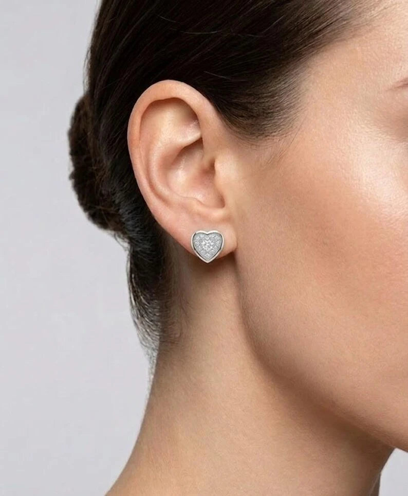 A close-up of a model's ear wearing a small heart-shaped stud earring with a silver-toned border and a clear central stone.
