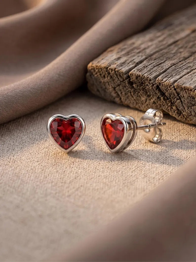 A pair of Red heart stud earrings ,  with faceted deep red gemstones in polished silver settings, displayed on a textured beige fabric next to a rustic wooden block.