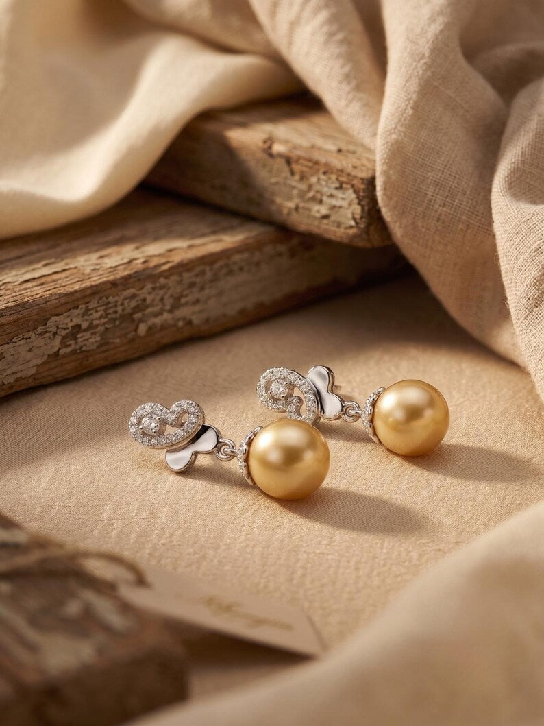 A pair of gold and silver drop earrings with heart-shaped crystal clusters and round gold pearl drops, displayed on a beige textured surface with draped fabric and rustic wood in the background.
