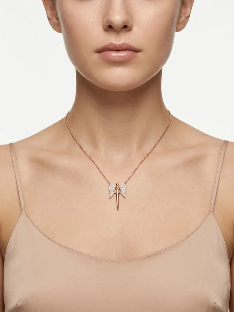 A close-up of a model wearing a delicate rose gold necklace with a bow-shaped pendant adorned with small clear crystals, paired with a beige camisole top against a light gray background.