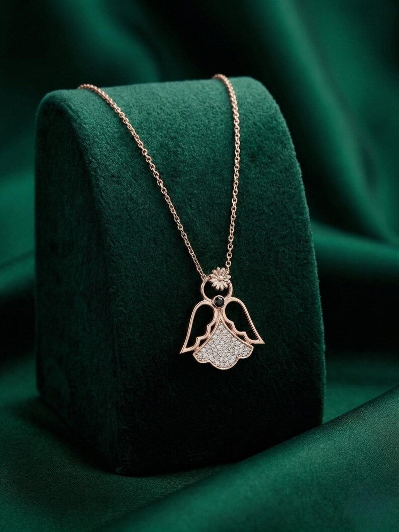 A rose gold angel pendant necklace with a crystal-encrusted lower wing, displayed on a dark green velvet jewelry stand against a matching green fabric background.