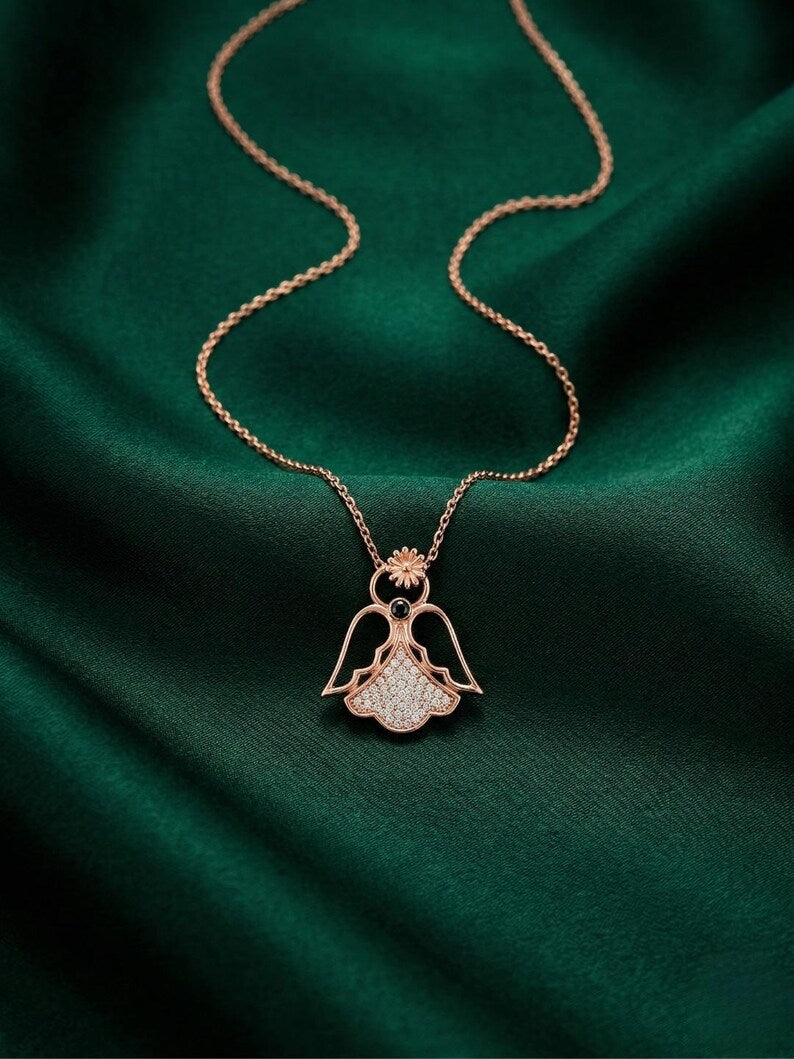 A rose gold angel pendant necklace with a crystal-encrusted body, resting on draped dark green satin fabric.