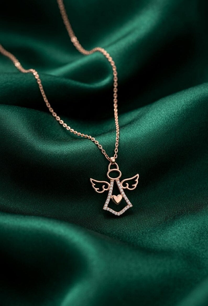 A rose gold angel pendant necklace with a small heart at the center, resting on a draped dark green satin fabric background.