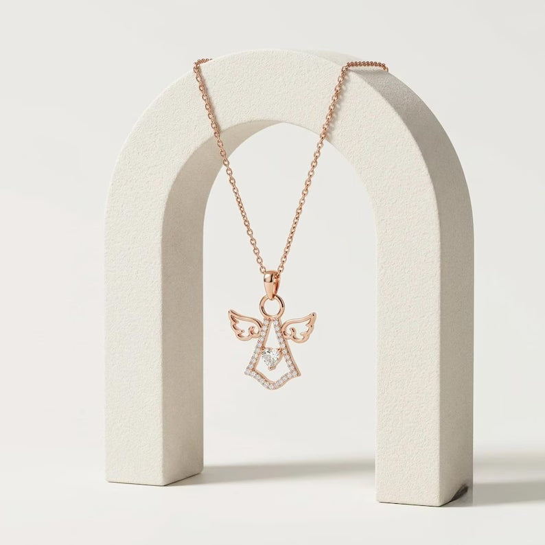 A rose gold angel pendant necklace with small clear stones, displayed on a textured white arch stand against a light gray background.