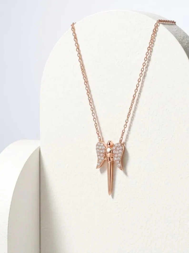 A rose gold angel pendant necklace with wing details paved with small clear stones, displayed on a white curved jewelry stand.