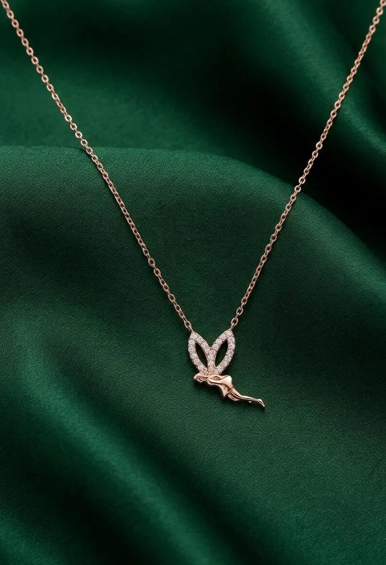 A delicate rose gold chain necklace featuring a small fairy pendant with sparkling clear stones, displayed on a draped dark green satin fabric background.