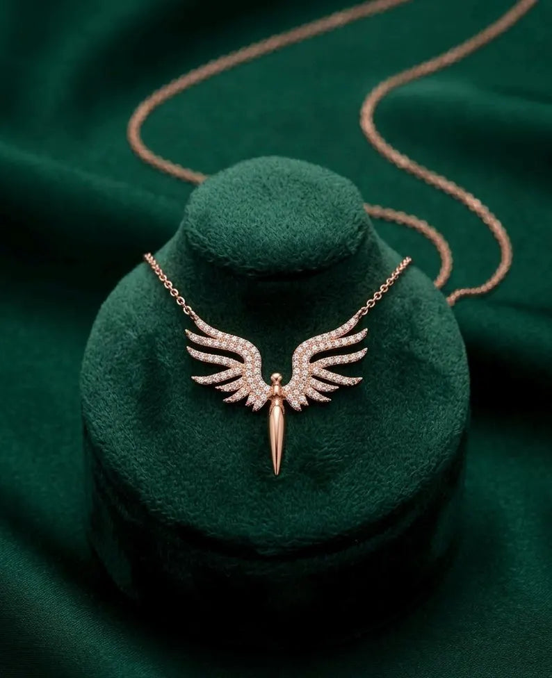 A rose gold angel wing pendant necklace with small clear stones, displayed on a dark green velvet jewelry bust against a matching dark green fabric background.