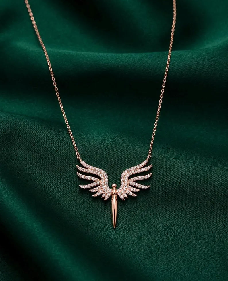 A rose gold angel wing pendant necklace with small clear pavé stones on the wings and a pointed dagger-hilt design hanging below, displayed on a draped dark green satin fabric background.