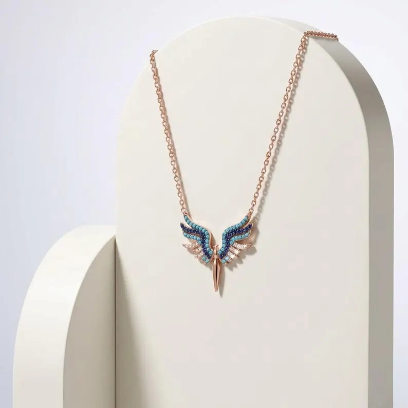 A rose gold necklace with a winged pendant featuring blue and white textured details, displayed on a white curved stand against a light gray background.