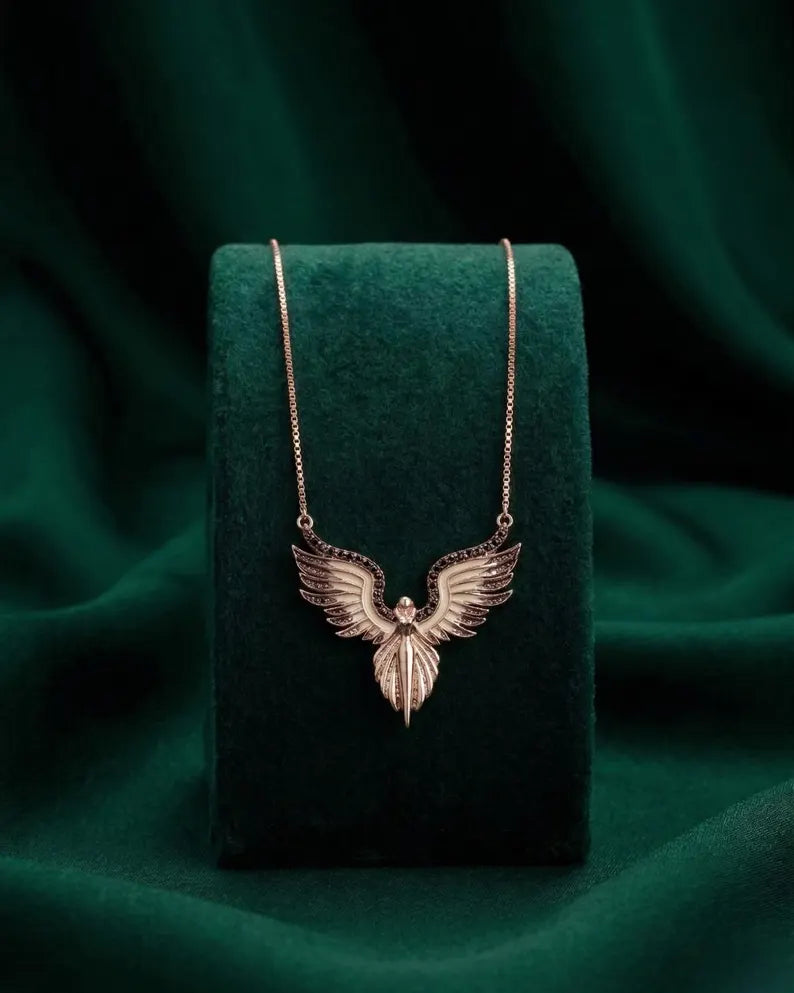 A rose gold winged pendant necklace displayed on a dark green velvet jewelry stand against a matching dark green draped fabric background.