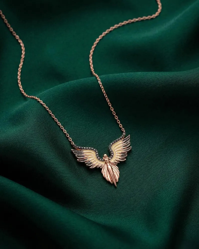 A rose gold angel pendant necklace with outstretched wings, resting on draped dark green satin fabric.