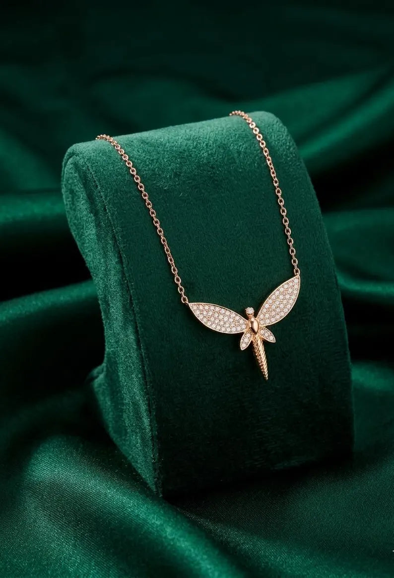 A rose gold necklace with a dragonfly pendant paved with small clear stones, displayed on a dark green velvet jewelry stand against a matching green satin background.
