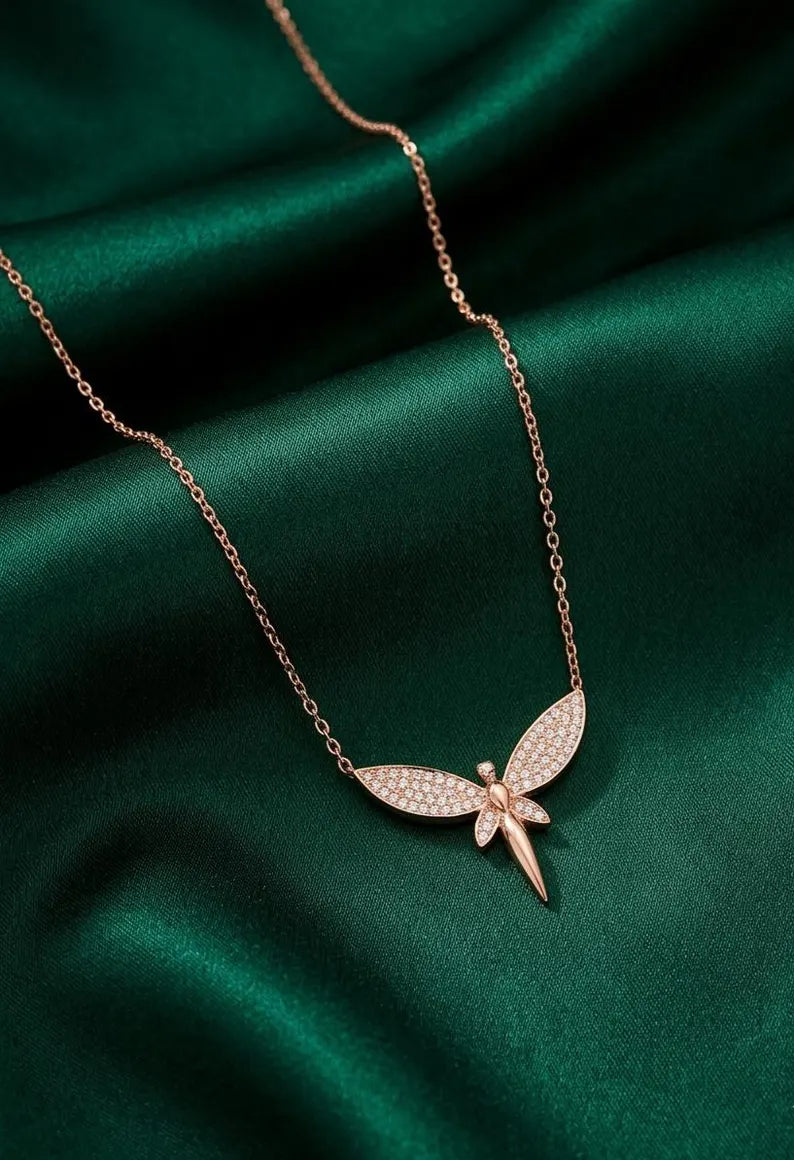 A rose gold dragonfly pendant necklace with pavé-set clear stones, displayed on a draped dark green satin fabric background.