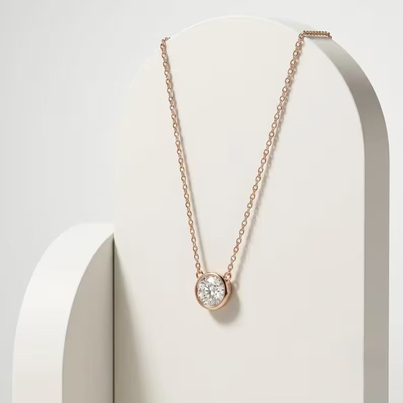 A delicate rose gold chain necklace with a round bezel-set clear gemstone pendant, displayed on a minimalist white geometric stand.