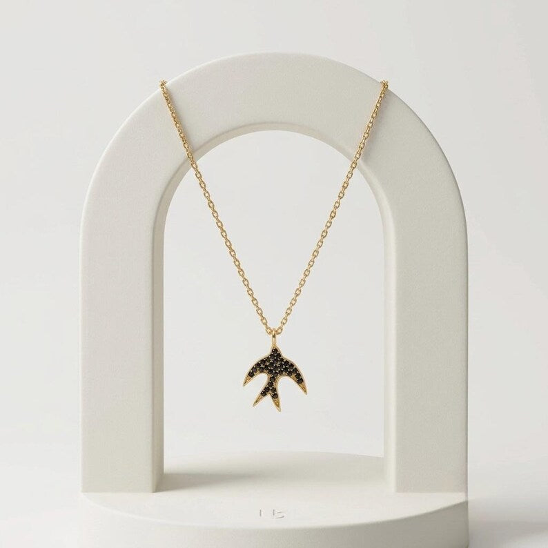 A gold chain necklace with a swallow-shaped pendant studded with small black stones, displayed on a white arch stand against a light gray background.
