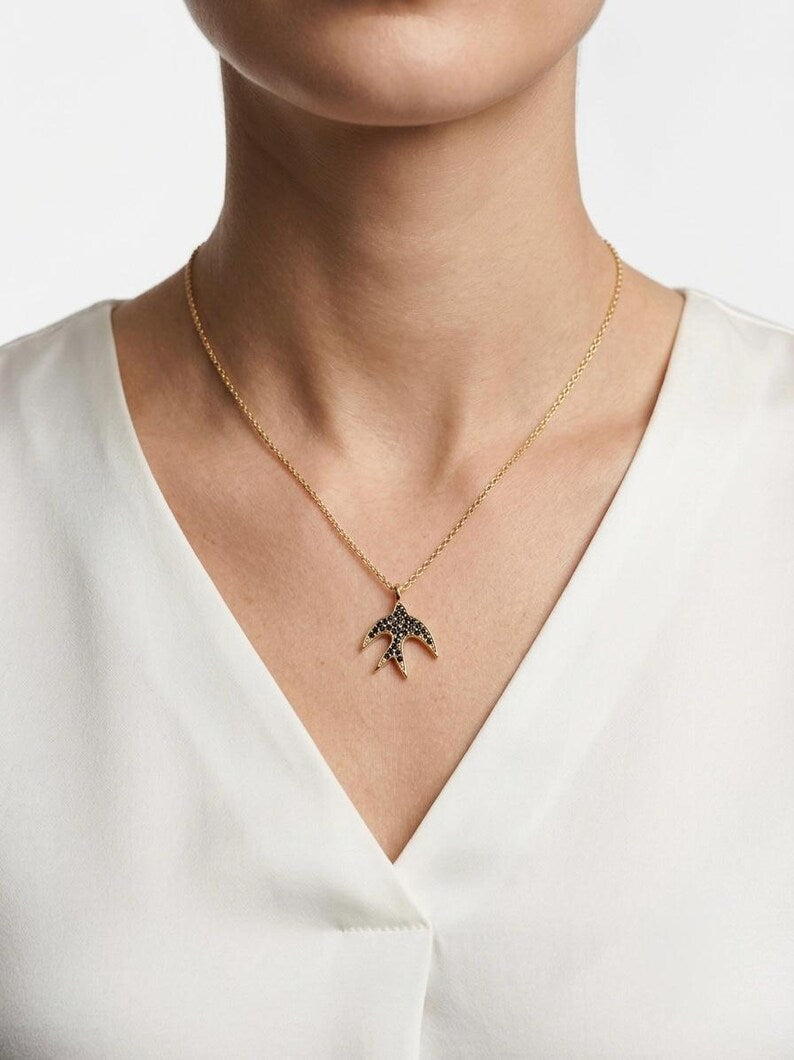 A gold chain necklace with a bird-shaped pendant embellished with small dark stones, worn by a model in a white V-neck top against a light gray background.