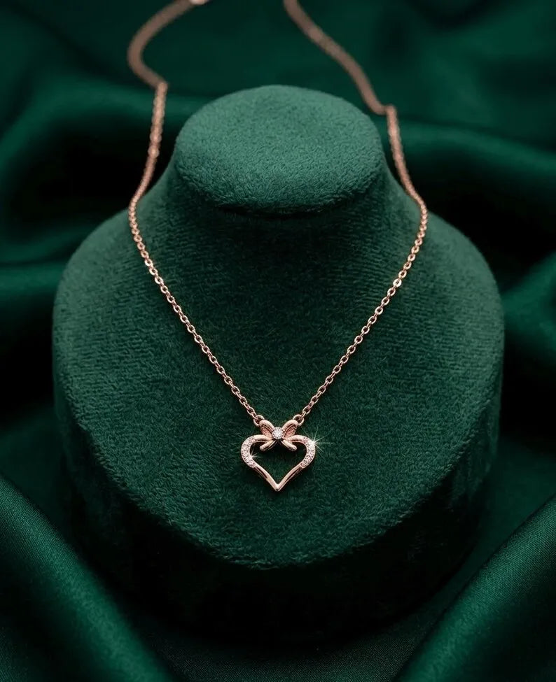 A delicate rose gold necklace with a heart-shaped pendant displayed on a dark green velvet jewelry bust against a matching green satin background.