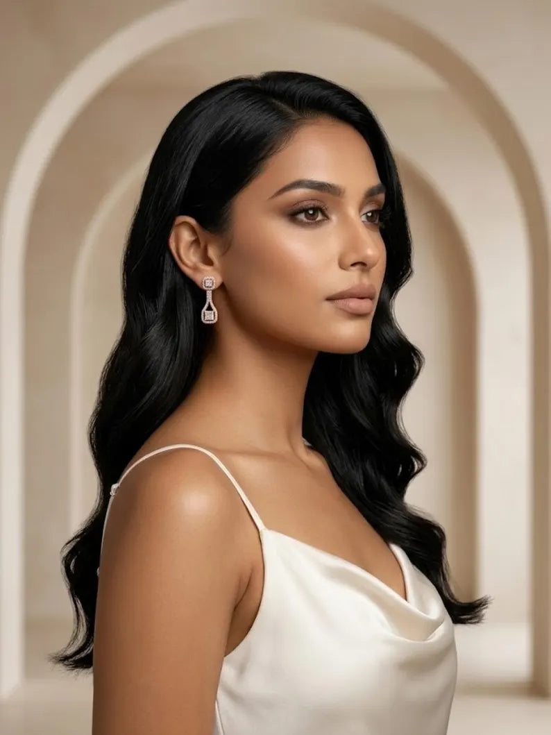 A woman with long, wavy black hair is shown in profile wearing a white spaghetti-strap top and a dangling silver-toned earring with a clear teardrop-shaped stone. She is photographed against a softly blurred beige architectural background with arches.