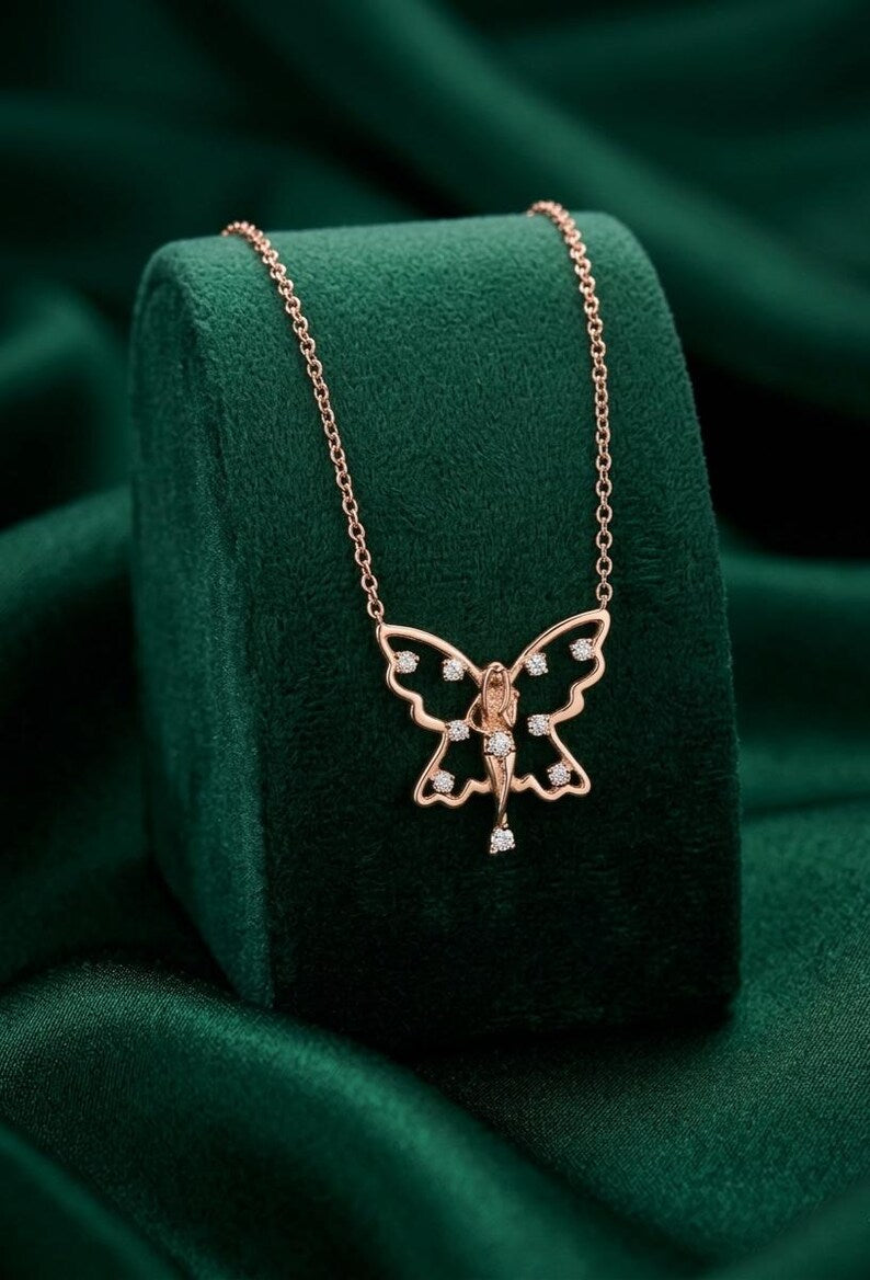 A rose gold fairy necklace with a delicate winged pendant and small clear stones, displayed on a dark green velvet jewelry stand against a matching green fabric background.