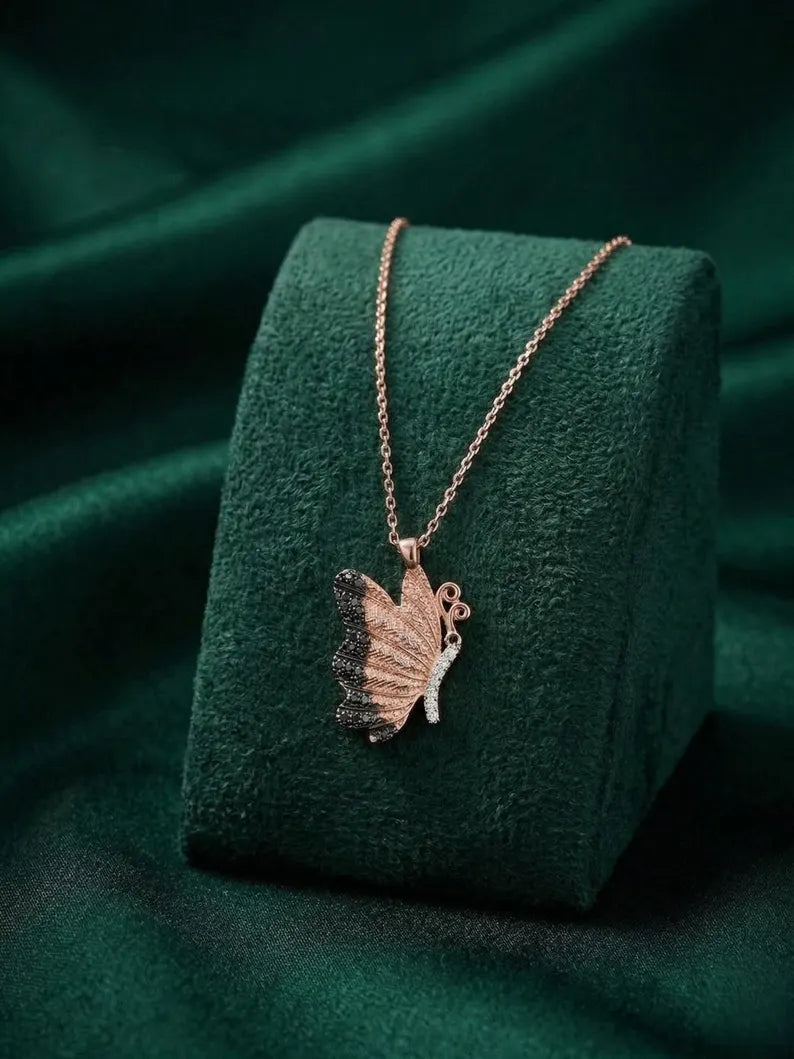 A rose gold butterfly pendant necklace displayed on a dark green velvet jewelry stand against a matching dark green fabric background.
