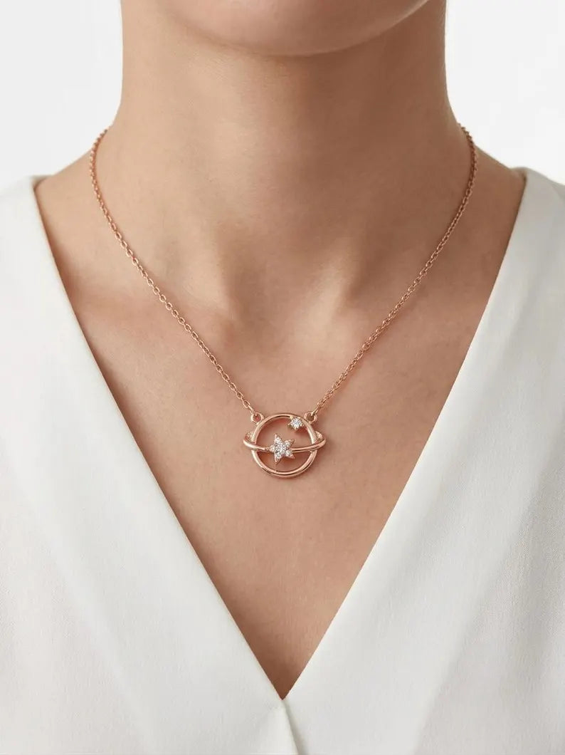 A close-up of a model wearing a delicate rose gold necklace with a circular orbit and star pendant, styled with a white V-neck top.