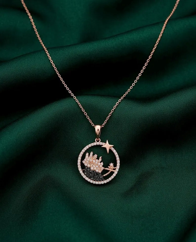 A rose gold necklace with a circular pendant featuring a crescent moon, star, and a small castle, displayed on a dark green satin fabric background.