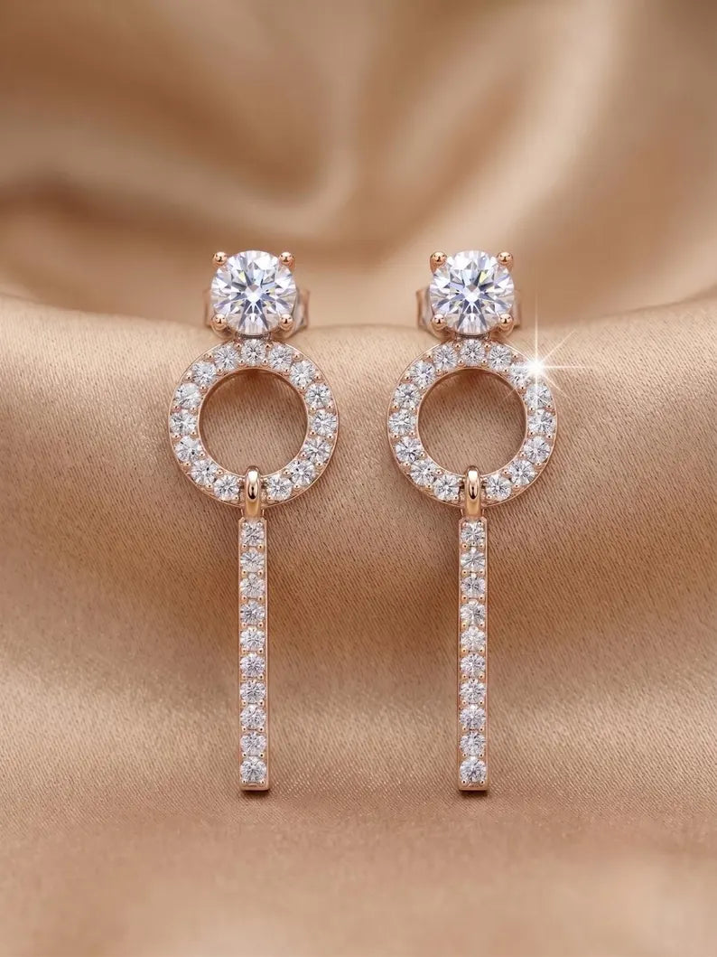 A pair of rose gold dangle earrings with clear cubic zirconia stones, displayed on a draped beige satin fabric background.
