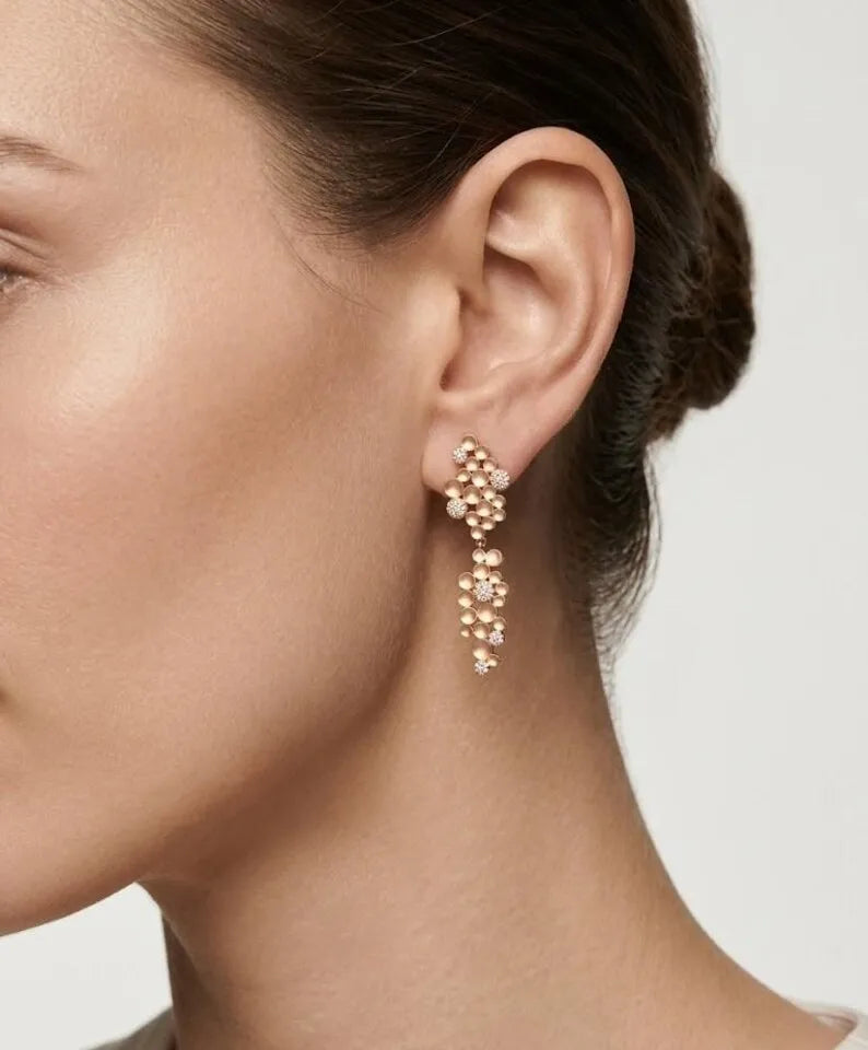 A close-up of a model's ear wearing a delicate gold dangle earring adorned with small clustered pearls and clear rhinestones, set against a clean light gray background.