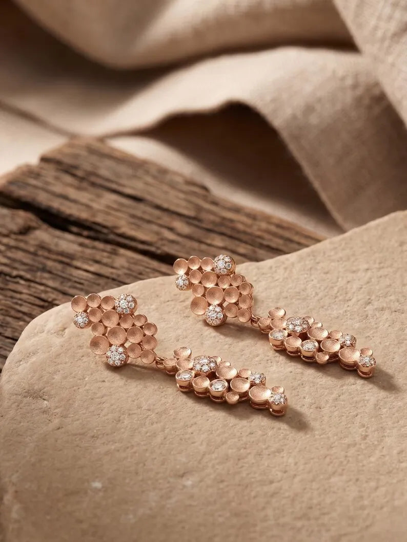 Rose gold dangle earrings with textured floral clusters and small clear stones, displayed on a beige stone surface with a rustic wooden prop and draped fabric in the background.