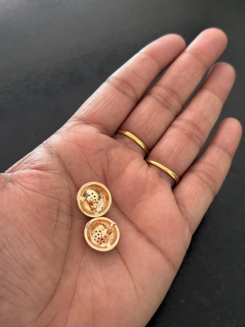 A close-up of a person's open palm holding two small gold round jewelry pieces, with two matching gold rings worn on the fingers, against a dark background.