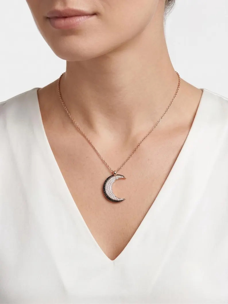 A close-up of a model wearing a delicate rose gold crescent moon pendant necklace with a row of small clear stones, paired with a white V-neck top.