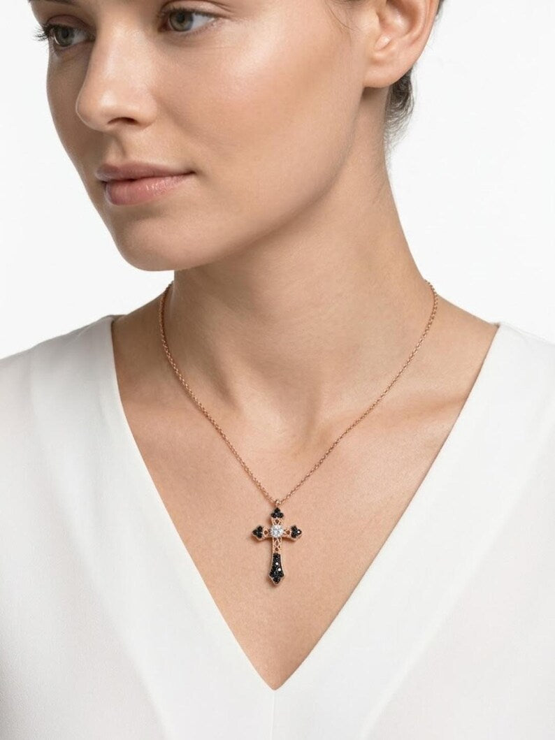 A close-up of a woman wearing a delicate rose gold cross pendant necklace with dark blue gemstone accents, paired with a white V-neck top against a clean white background.