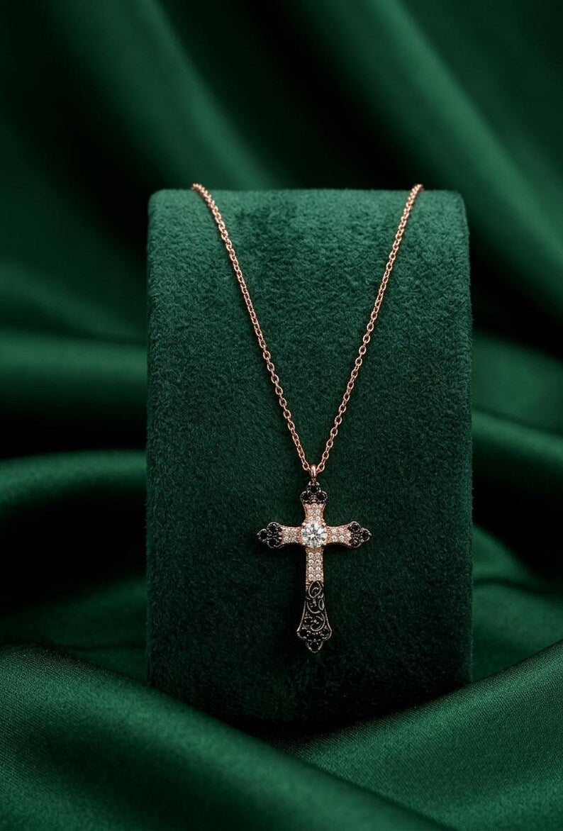 A rose gold cross pendant necklace with small clear stones, displayed on a dark green velvet jewelry stand against a draped green satin background.