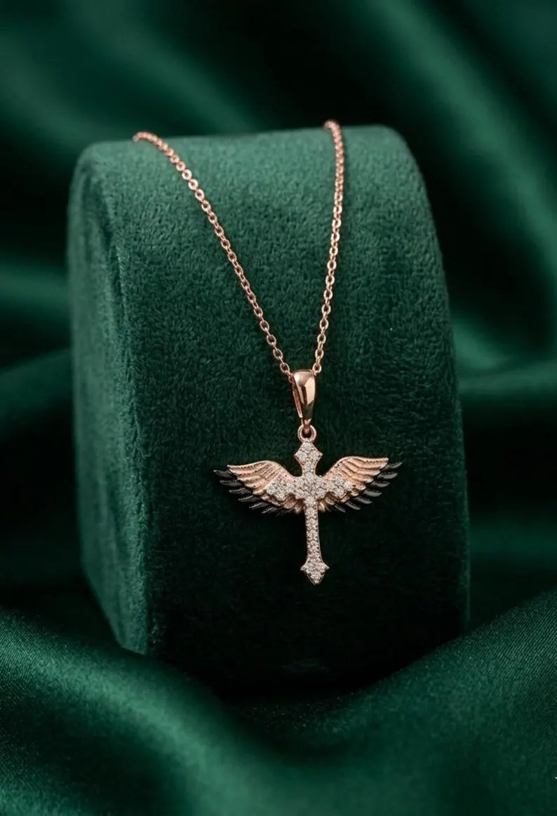 A rose gold cross pendant necklace with wing details and small clear stones, displayed on a dark green velvet jewelry stand against a matching green fabric background.
