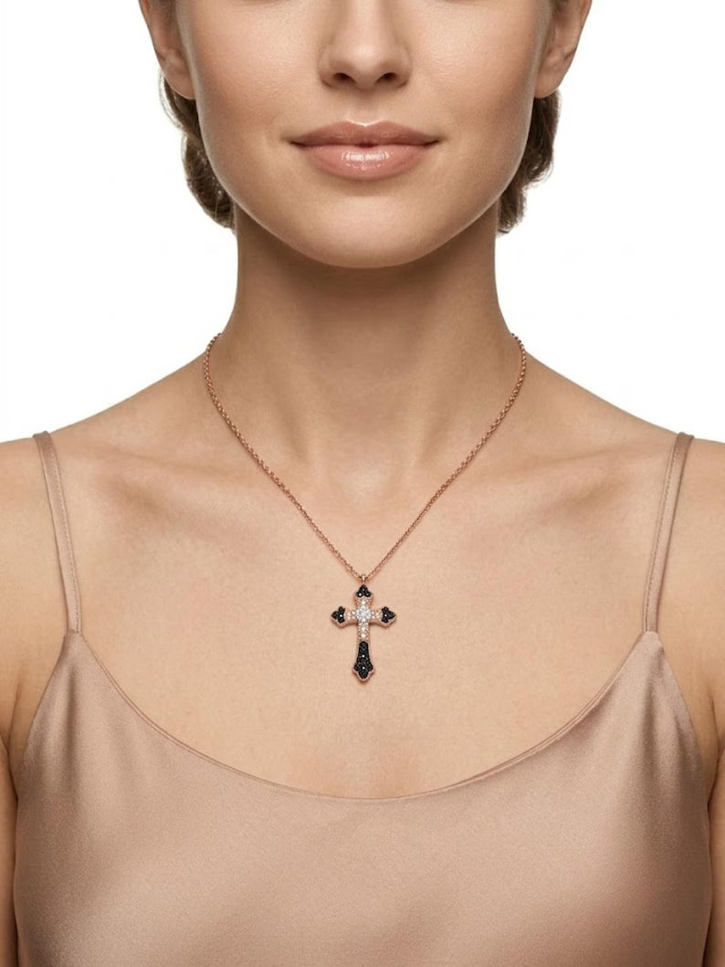 A close-up of a model wearing a delicate rose gold cross pendant necklace with dark blue and clear stones, paired with a beige satin camisole top against a white background.