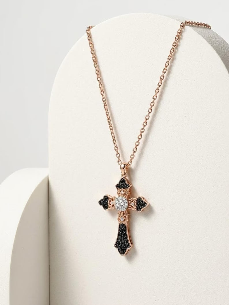 A rose gold cross pendant necklace with black pavé accents and a clear central gemstone, displayed on a cream-colored curved stand.